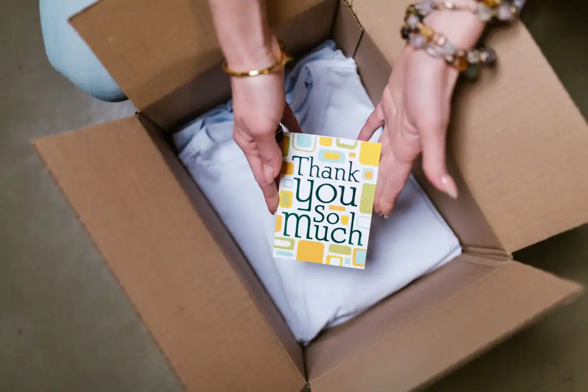 a person holds a thank you card above items in an open shipping box, suggesting care in sending.
